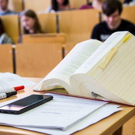 Aufgeschlagenes Buch vor Sitzreihe mit Studierenden.