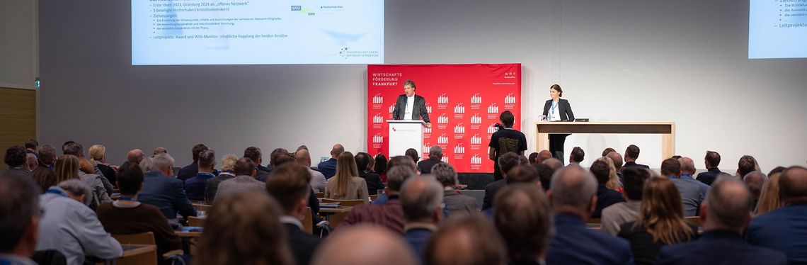 Großer Konferenzraum mit einer Rednerin und einem Redner und Publikum.