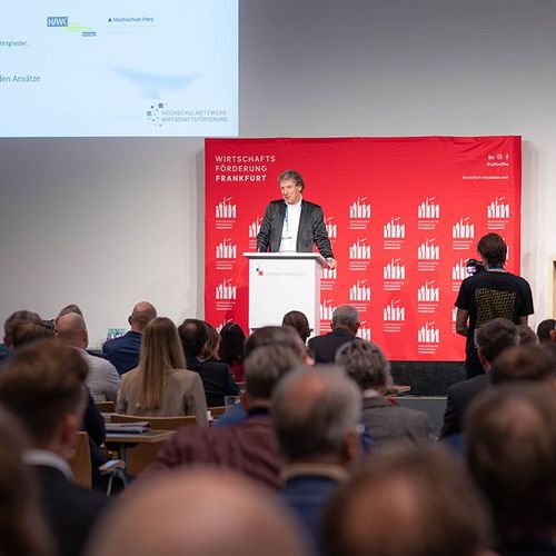 Großer Konferenzraum mit einer Rednerin und einem Redner und Publikum.