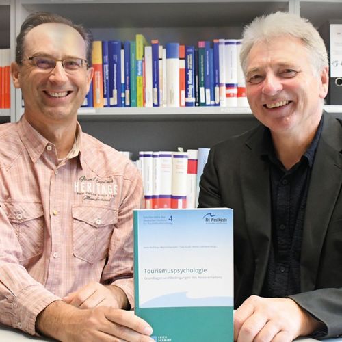 Zwei Männer sitzen an einem Tisch, sie halten ein Buch in den Händen. Im Hintergrund ist ein Bücherregal zu sehen.