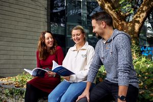 Drei Studierende des Masterstudiengangs Public Management sitzen lachend auf einer Bank unter einem großen Baum und haben einige Bücher in der Hand.