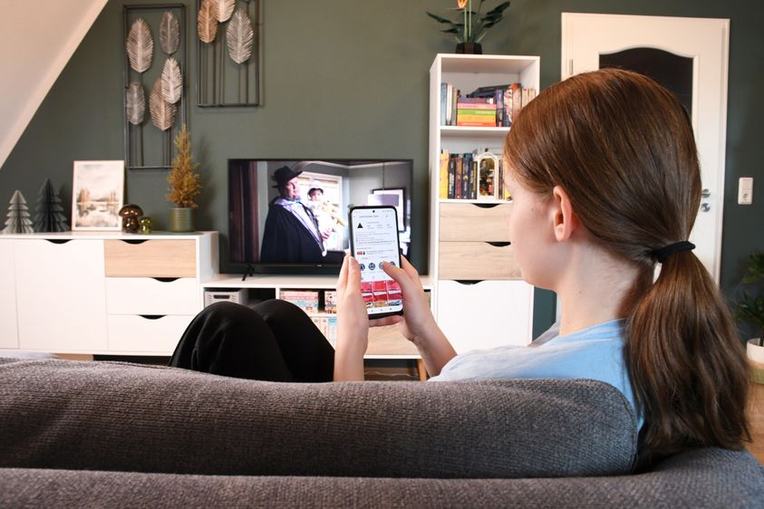 Ein Teenager-Mädchen sitzt vor dem laufenden Fernseher, sie bedient parallel ein Smartphone.