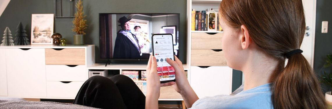 Ein Teenager-Mädchen sitzt vor dem laufenden Fernseher, sie bedient parallel ein Smartphone.