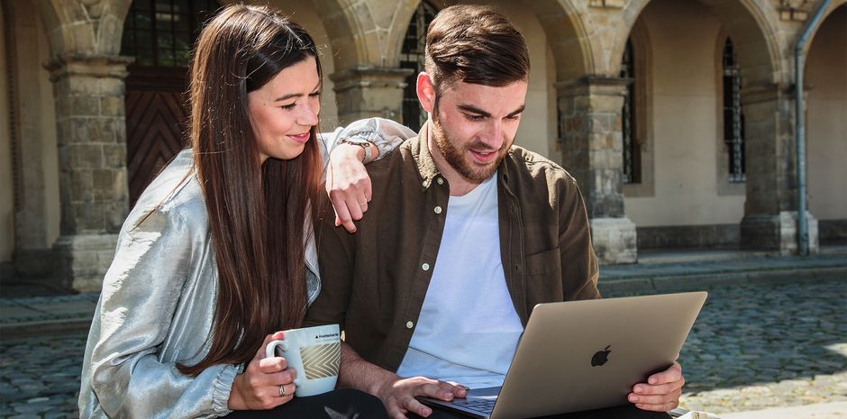 Eine Studentin und ein Student des IT-Management - Verwaltungsinformatik auf dem Domplatz vor den Gebäuden des Fachbereichs Verwaltungswissenschaften schauen gemeinsam auf einen Laptop.