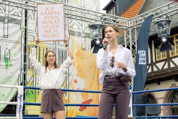 Zwei Frauen im Boxring, eine spricht ins Mikrofon, eine hält ein Plakat mit der Aufschrift "Ich kämpfe jeden Monat gegen mich selbst"..