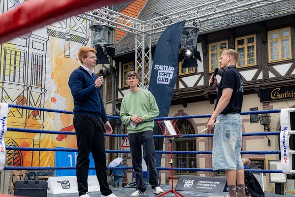 Drei Männer im Boxring, einer spricht ins Mikrofon, in der Mitte steht ein Notenständer