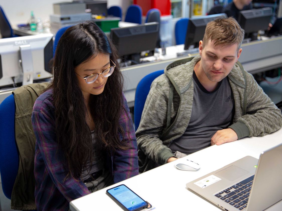 Symbolbild CyberSecurity Forschung: Zwei junge Menschen sitzen an einem Tisch und sehen auf ein Smartphone