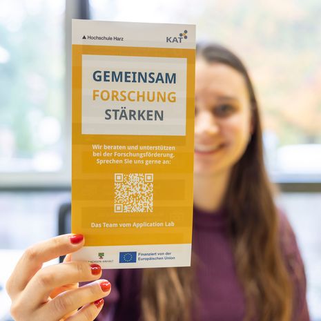Eine Frau hält einen Flyer in die Kamera, auf dem steht "Gemeinsam Forschung stärken"