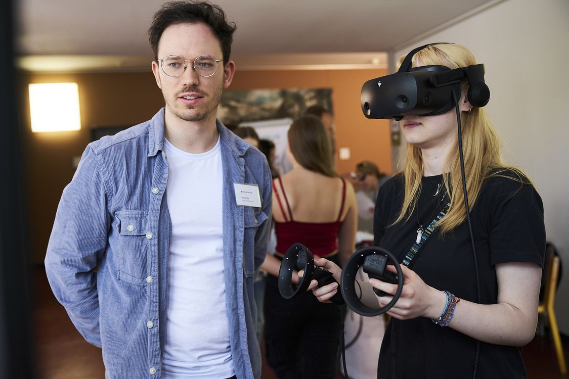 Ein Hochschulmitarbeiter in lockerer Alltagskleidung zeigt einer Studentin die Einsatzmöglichkeiten von Virtual Reality. Sie trägt eine VR-Brille und hält zwei Controller in Händern.